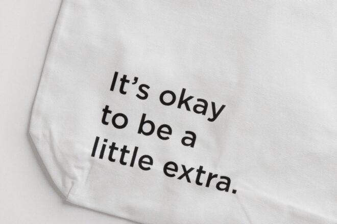 a white tote bag with black text that reads "It's okay to be a little extra."