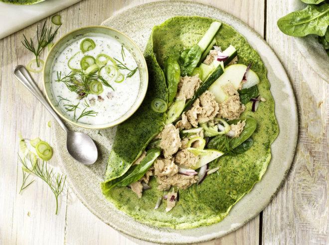 tuna salad on a green tortilla, next to a bowl of white sauce with green vegetables sliced on top