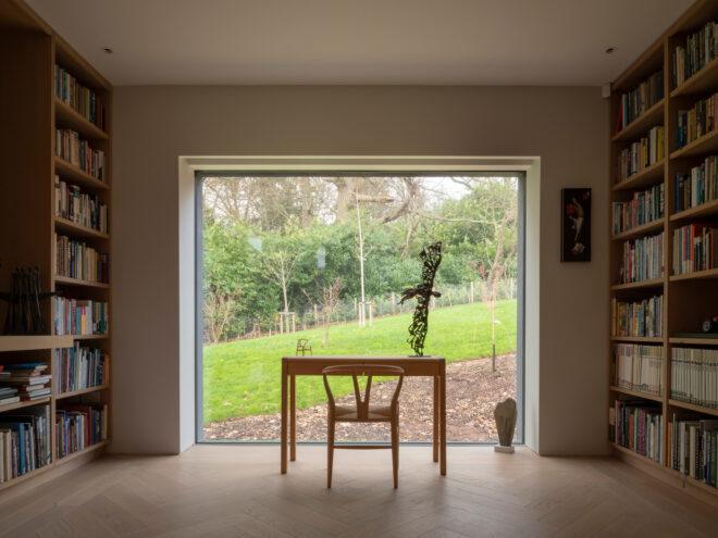 wood desk facing large window in room with two walls of floor-to-ceiling bookshelves