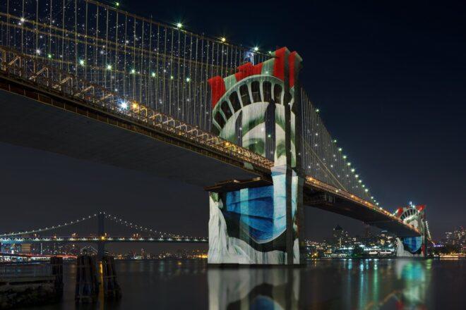 photo of masked Lady Liberty image projected onto the Brooklyn Bridge