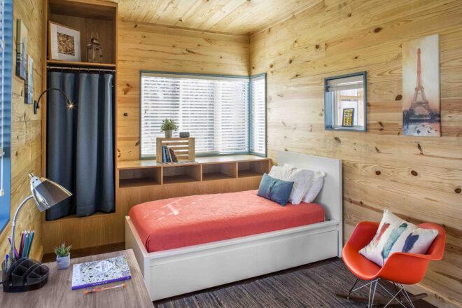 a bedroom with wood walls, patterned gray flooring, a desk with lamp, an orange-red chair, and a white bed with a red bedspread.