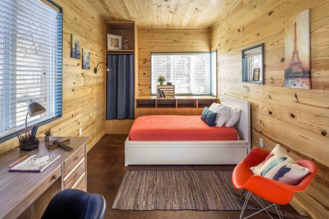 a bedroom with wood walls, patterned gray flooring, a desk with lamp, an orange-red chair, and a white bed with a red bedspread.