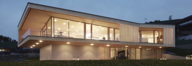 a two-story home. the lower floor features an opaque white facade on the left side, and glazing on the right side. the upper floor features wraparoun glazing framed by an overhanging timber facade. interior lights make the home glow.