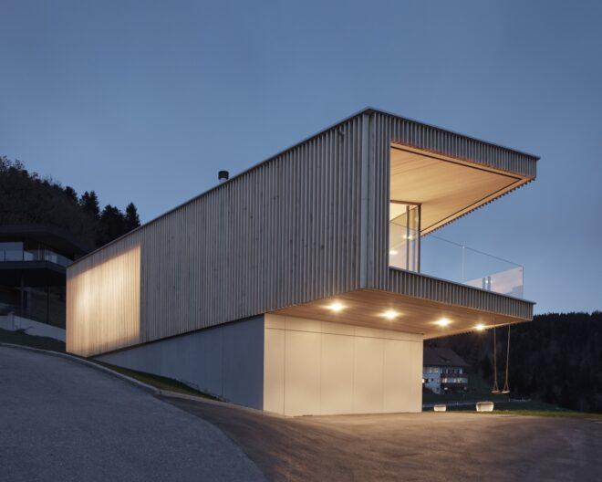 a C-shaped structure with a facade of vertical timber slats rests atop a solid lower story with a white facade. bright LED lights illuminate the home. a swing hangs from the second floor overhang.