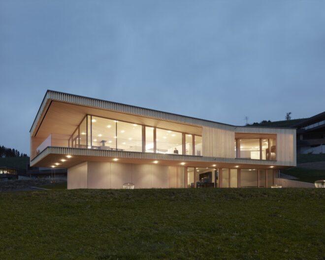 a two-story home. the lower floor features an opaque white facade on the left side, and glazing on the right side. the upper floor features wraparoun glazing framed by an overhanging timber facade. interior lights make the home glow.