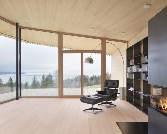 an interior living space with wood floors and glazed walls that look out onto a forest and lake. a floor lamp hangs over a dark leather-look chair and foot stool. on the right, a black bookshelf stands against the wall next to a fireplace.