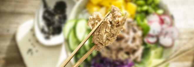 a pair of chopsticks holding flakes of tuna against a blurry background of a bowl of tuna salad