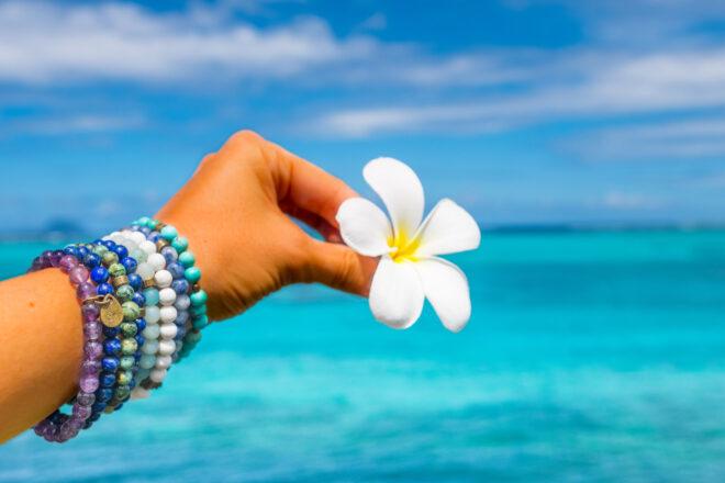 person wearing blue beaded bracelets and holding tropical flower with ocean in the background
