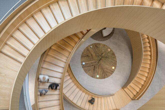 an overhead view of a spiral staircase made of light-colored wood. blurry people walk down the staircase.