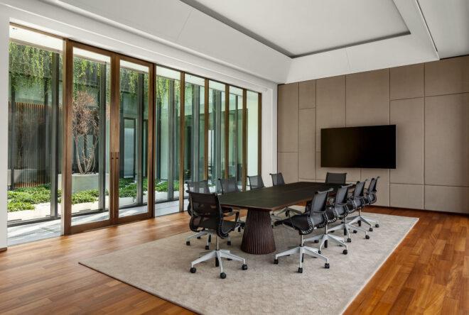 a meeting room with a long, dark wood table surrounded by 10 black and white office chairs. a TV hangs on the wall toward the end of the conference table. to the left are doorways and floor-to-celing windows.