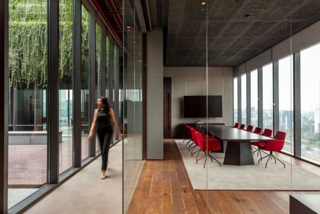 to the left, a hallway with a blurry person walking through and looking out the floor-to-ceiling windows. to the right, a conference room with glass walls and a meeting table surrounded by red chairs. a TV hangs on the wall toward the end of the table.