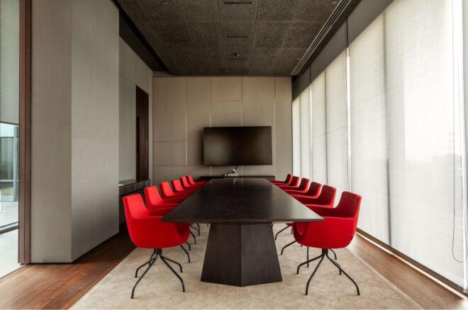 a conference room with glass walls on the right, covered by sheer white curtains, and a meeting table surrounded by red chairs. a TV hangs on the wall toward the end of the table.