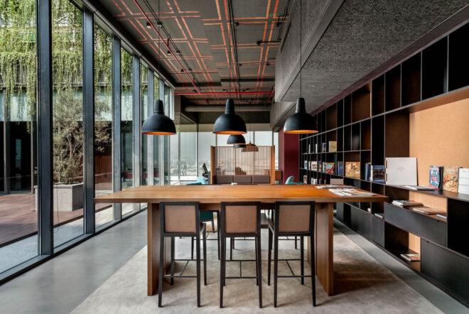 an office space with floor-to-ceiling windows to the right and a black bookshelf built in to the wall on the right. in the middle of the room is a wood table with three tall chairs and three black lamps hanging over the top of the table.