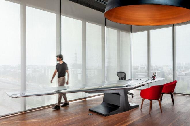 an office surrounded by floor-to-ceiling windows covered in sheer white curtains. a blurry person walks behind a large desk made from a recycled airplane wing. there is a black office chair on one side of the desk and two red chairs on the opposite side of the desk.