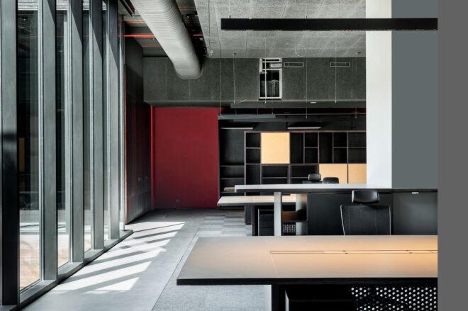 an industrial-style office space with part of the wall painted red. to the left are floor-to-ceiling windows. various tables and chairs fill the room.