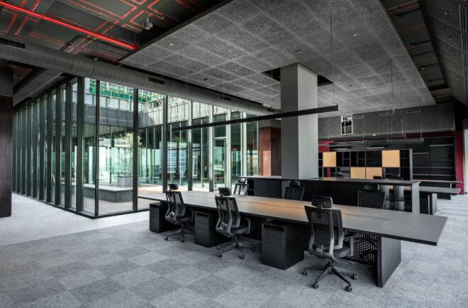 an office space with floor-to-ceiling windows. the room features gray carpeting and long tables surrounded by office chairs.
