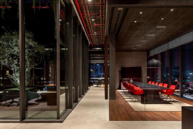 to the left, a hallway with floor-to-ceiling windows. to the right, a conference room with glass walls and a meeting table surrounded by red chairs. a TV hangs on the wall toward the end of the table. out the windows you can see it is dark outside.