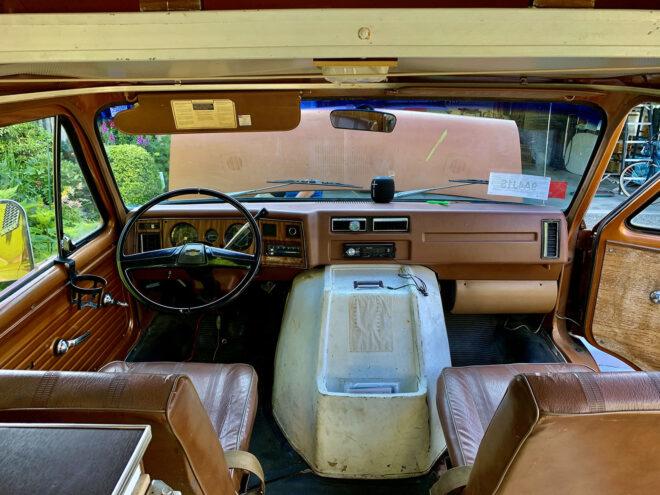 the front of a brown and off-white 1975 Chevy van, showing the driver and passenger seats. through the windshield you can see the hood of the car raised.