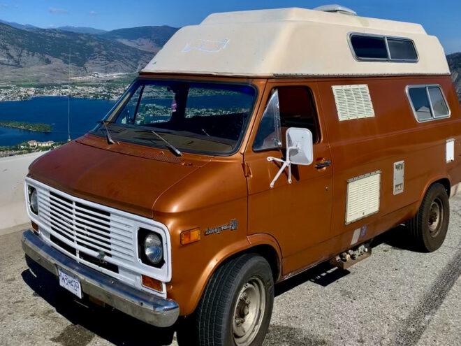 a brown and off-white 1975 Chevy van parked on the side of the road. in the background there is blue water and mountains.