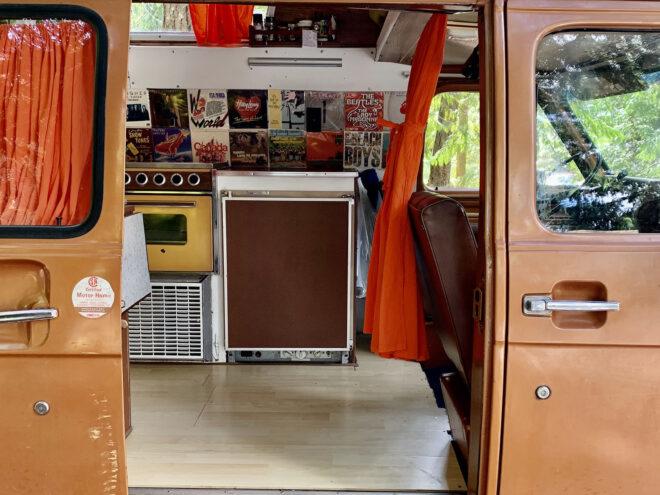 a brown and off-white 1975 Chevy with its sliding door opened to show a kitchen area with album covers as wall decorations.