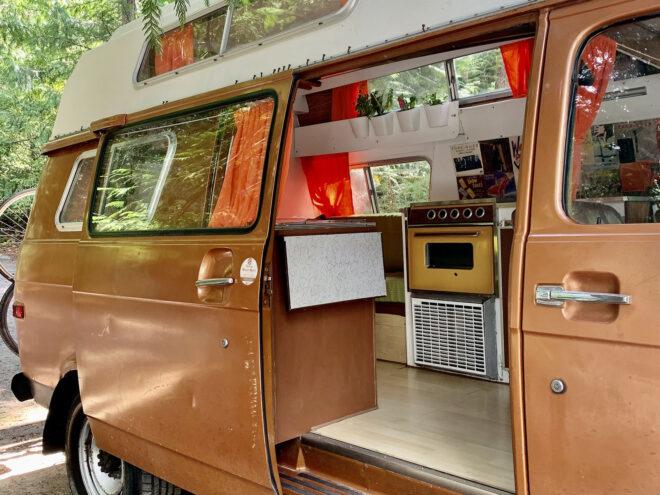 a brown and off-white 1975 Chevy with its sliding door opened to show a yellow stovetop/oven and indoor plants.