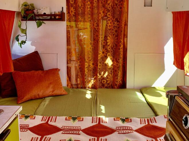 a foldout bed with green, white and red/orange sheets and a sheer orange curtain in the background.