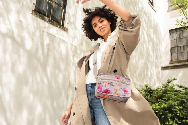 a person with dark, short hair wearing a beige jacket and raising one arm to show off a multi-colored (pink, orange and gray) purse with a crossbody strap.