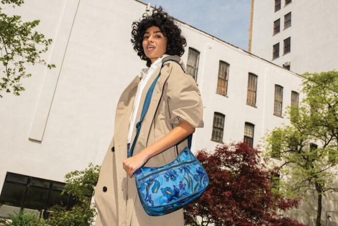 a person with dark, short hair wearing a beige jacket holding a blue purse that hangs off their shoulder via a long strap.
