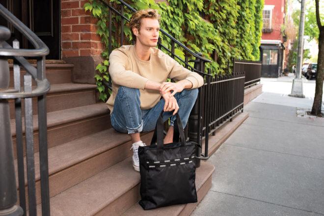 a person with short brown hair and a light-colored sweater sits on the stoop in fron of a brownstone, holding a black bag with a short strap.