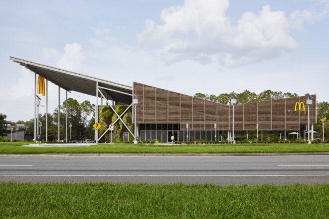 a streetview of a McDonald's restaurant with a sloped, v-shaped roof. a yellow "M" hangs from the edge of an overhang on the building, which features a facade of wood slats.