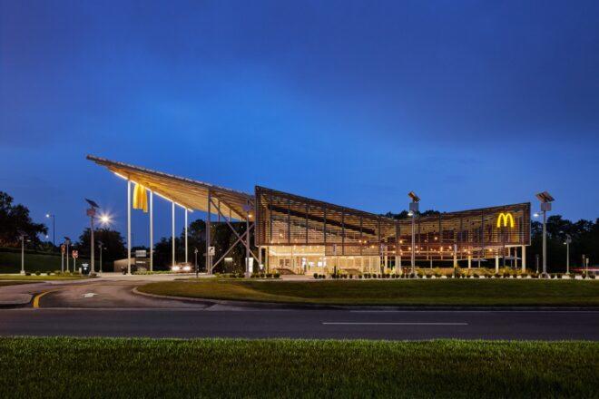 a night-time streetview of a McDonald's restaurant with a sloped, v-shaped roof. a yellow "M" hangs from the edge of an overhang on the building, which features a facade of wood slats.