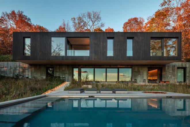 a multi-level home. the top floor has a dark timber facade with several windows of various sizes. the lower floor's facade uses concrete and features several windows. in front of the home is a pool area.