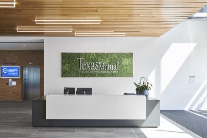 a lobby with elevators set into a brick wall to the left. to the right are white walls with a green in-set sign reading "Texas Mutual." A dark gray and white desk sits in front of the green sign.