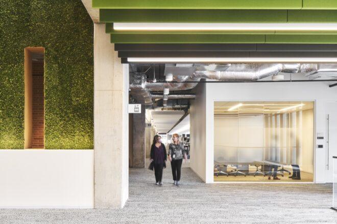 two people walking through a hallway with a greenery-covered wall on the left and a conference room on the right.
