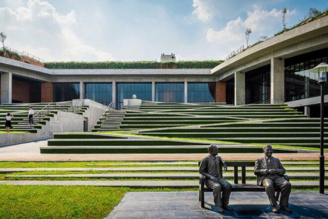 a terraced, green ampitheater area with two people on the left standing on the stairs. in the foreground to the right is a bench with two statues of men sitting on the bench.