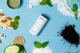 a white tube of deodorant against a blue background, surrounded by shredded coconut, mint leaves, activated charcoal powder, cucumber slices, nuts and a metal container of green liquid.