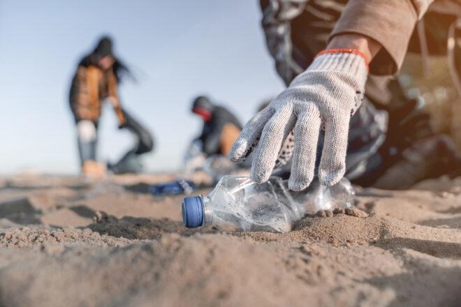 gloved hand picking up plastic bottle from sand