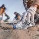 gloved hand picking up plastic bottle from sand