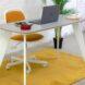 a white office desk with an orange and white desk chair with wheels. the desk has a laptop on it. underneath the table is a yellow rug.