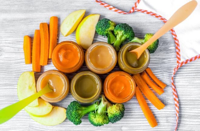 six jars of baby food in varying shades of orange. carrots, apples and broccoli surround the jars.