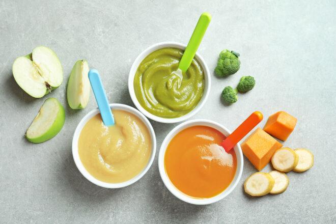 three bowls of baby food. from left to right, the contents of the jars are white, green and orange. apples, broccoli, squash and banana surround the bowls.