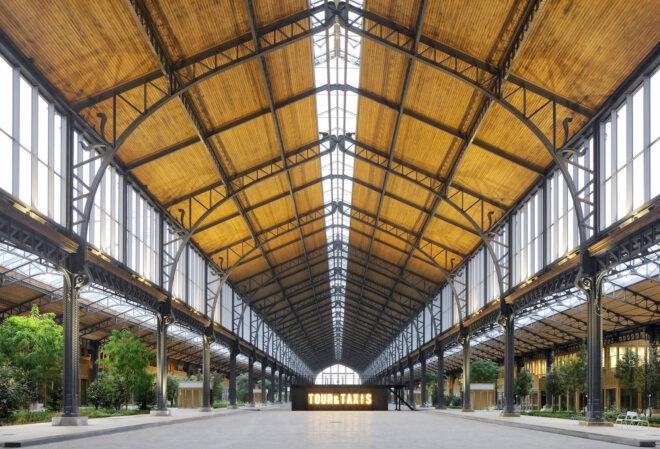 A dramatic, curved timber roof arching over an indoor shopping district. A sign in the center of the hall reads "Tour & Taxis."