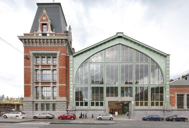 A tall, brick tower structure to the left, and a glass and green metal greenhouse-style building to the left.