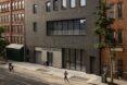 A city street with a boxy gray building that has a glass door entrance. People walk by in front of the building.