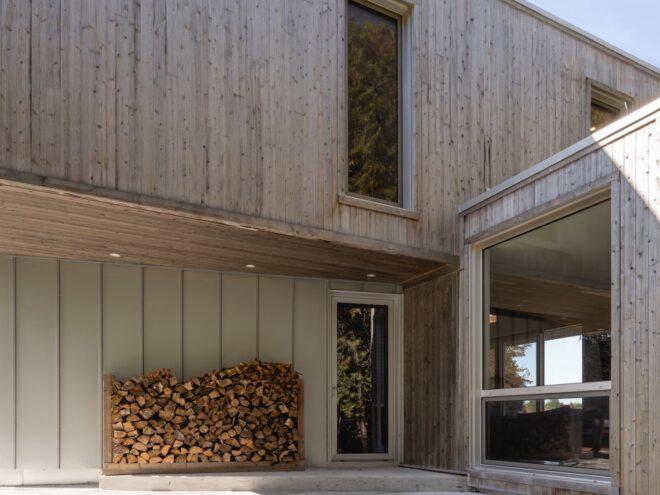 A two-story home with a gray facade broken up by large windows. The back of the home has a large pile of firewood.