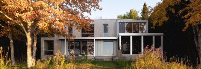 A two-story home with a gray facade broken up by large windows. The home is flanked on both sides with trees in fall colors.