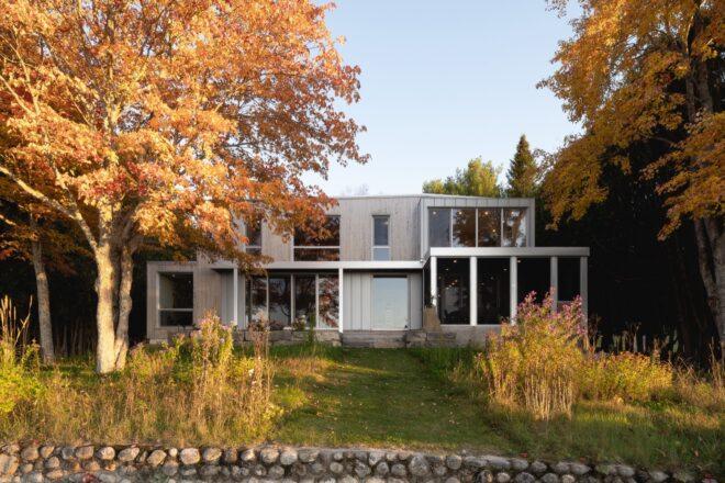 A two-story home with a gray facade broken up by large windows. The home is flanked on both sides with trees in fall colors.