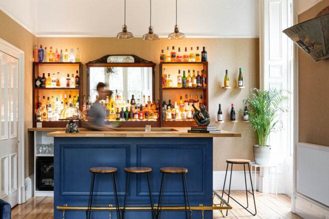 A bar area with a blue counter and three stools in front of it. Behind the counter, a blurry figure prepares drinks.
