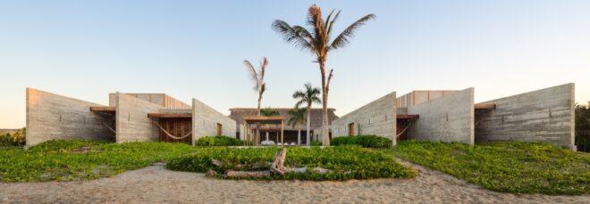The exterior of a beach house, with sand, greenery and palm trees in the front yard. The home is comprised of a U-shaped, off-white wood volume.