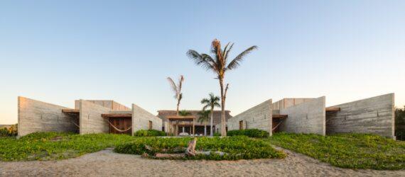 The exterior of a beach house, with sand, greenery and palm trees in the front yard. The home is comprised of a U-shaped, off-white wood volume.